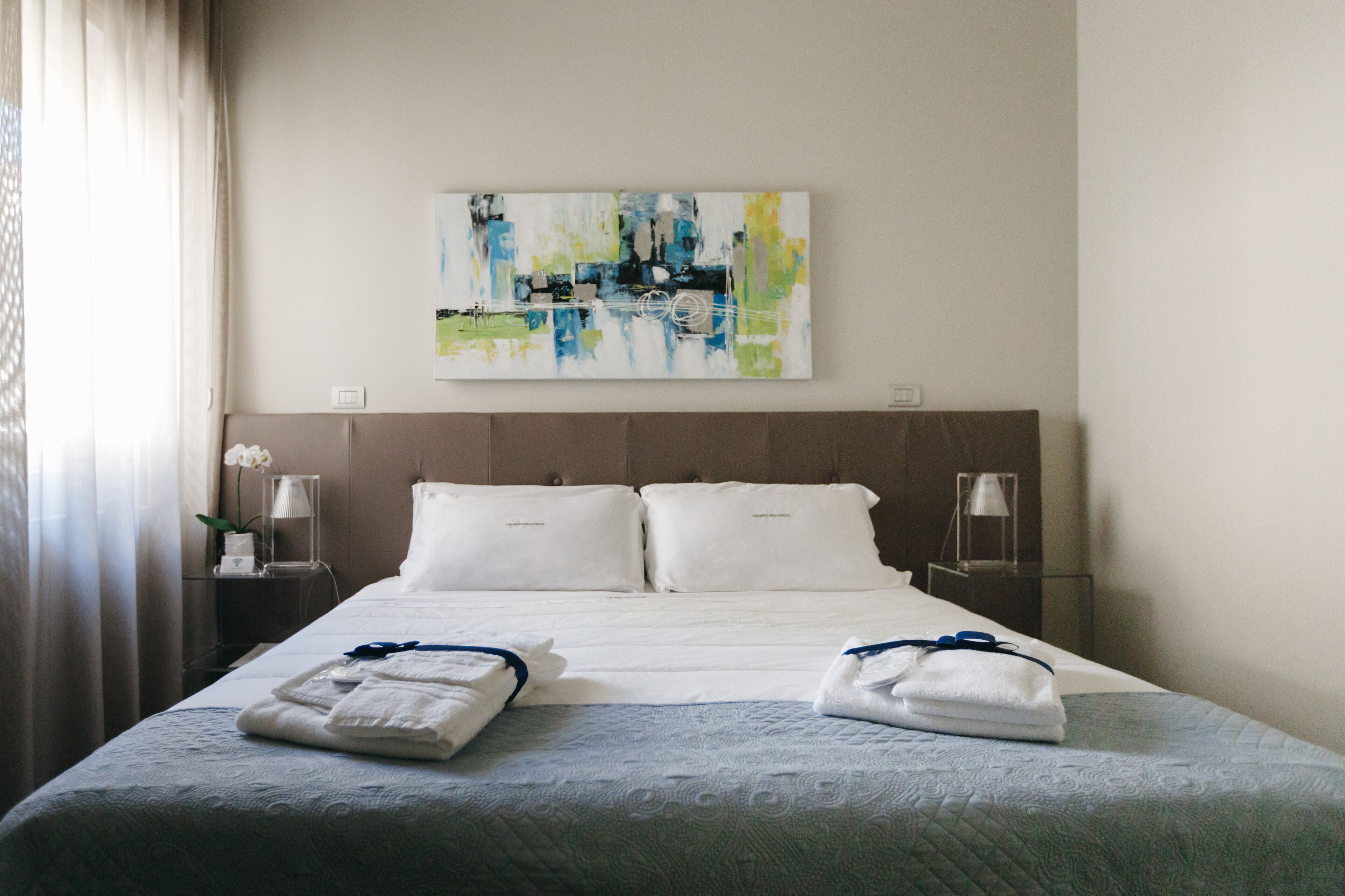 A hotel room with a large bed, two bedside tables, a wall-mounted abstract painting, and a window with curtains.