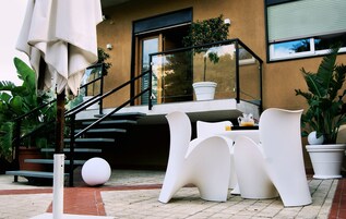 A modern outdoor seating area with white chairs and a table, a staircase leading to a balcony, and potted plants.