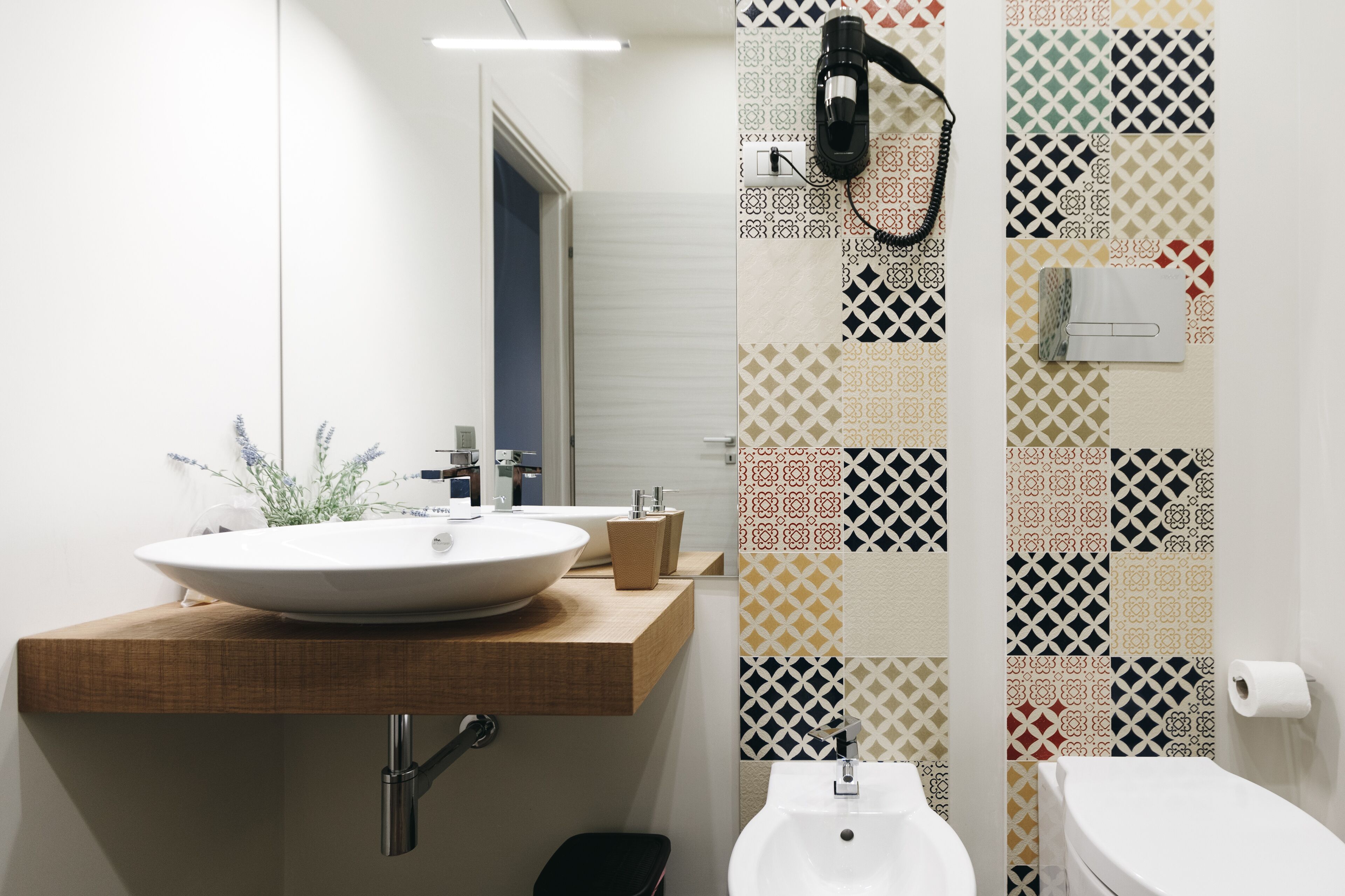 A modern bathroom with a large white sink, a wooden countertop, a mirror, and a patterned tile wall.