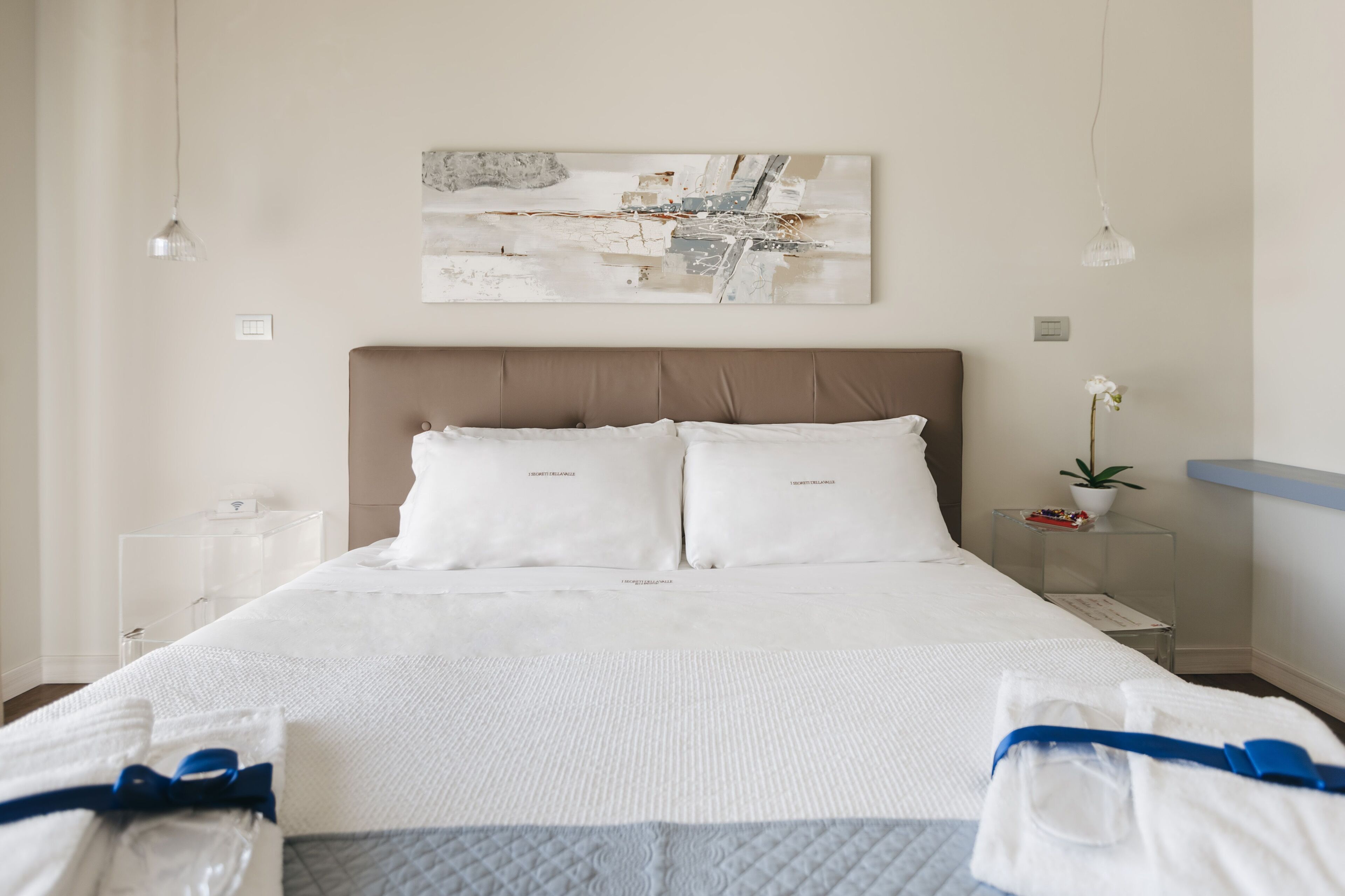 A neatly made bed with white linens, a modern headboard, and a minimalist decor including a wall art piece and a small potted plant.