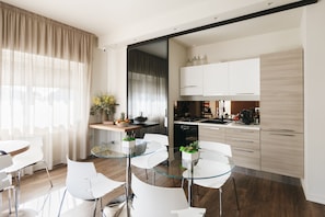 A modern kitchen with white cabinets, a black dishwasher, and a coffee machine. There is a dining area with a glass table and white chairs. - I Segreti della Valle (Agrigento)