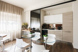 A modern kitchen with white cabinets, a black dishwasher, and a coffee machine. There is a dining area with a glass table and white chairs.