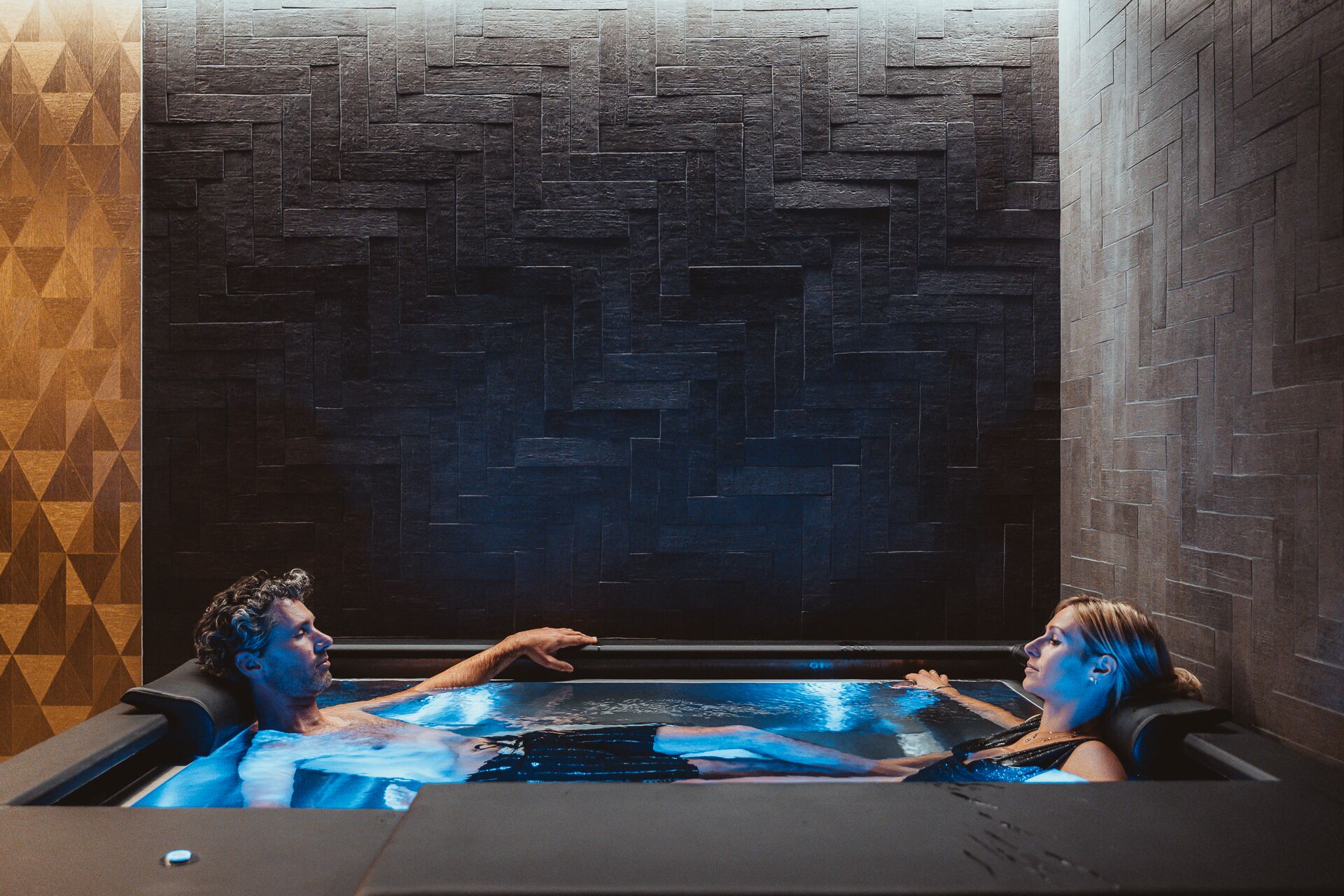 A hot tub with two people inside, surrounded by dark stone walls with a geometric pattern.
