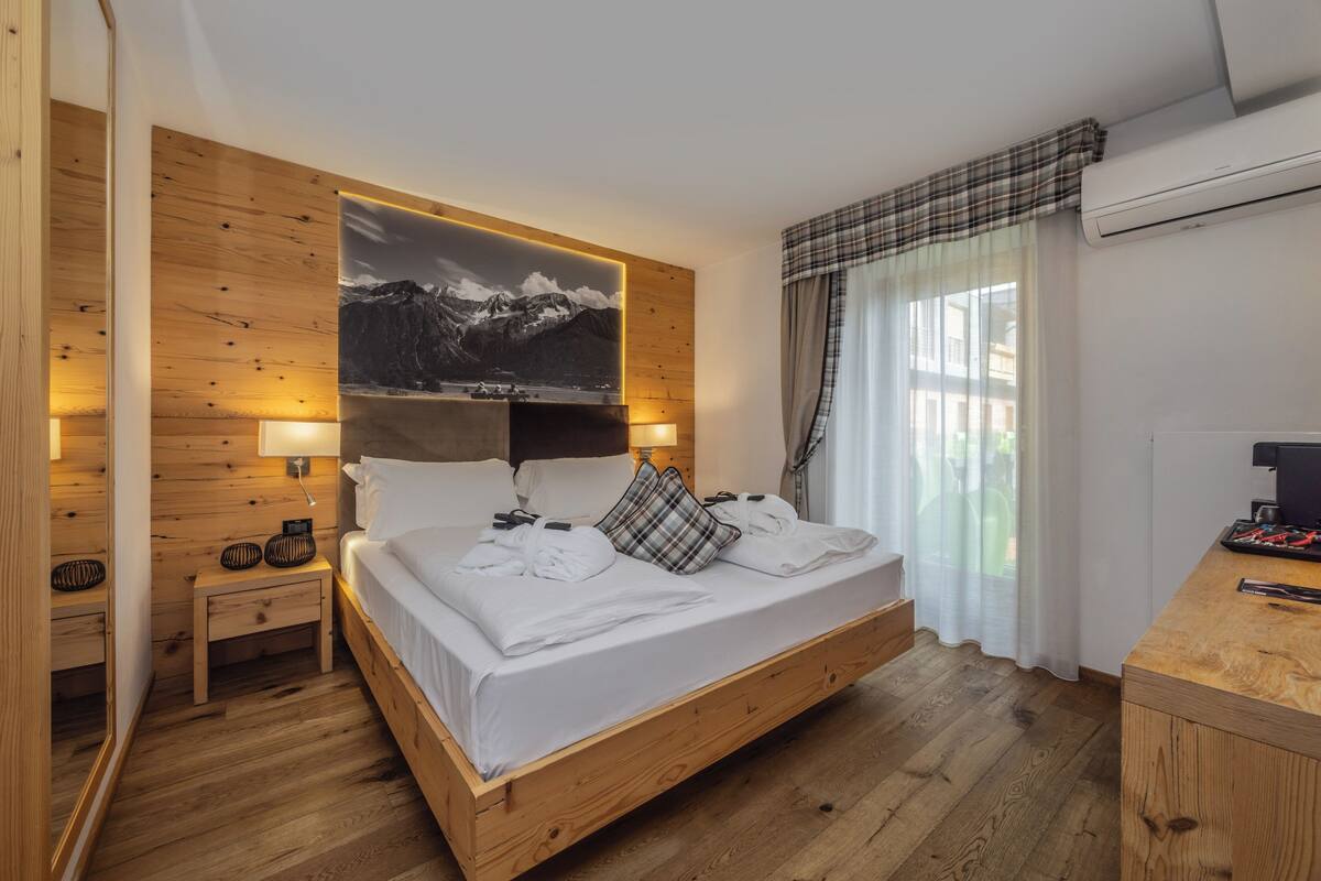 Un lit à cadre en bois, recouvert de draps blancs, avec un tableau représentant des montagnes accroché au mur et une table de chevet sur laquelle se trouve une lampe.