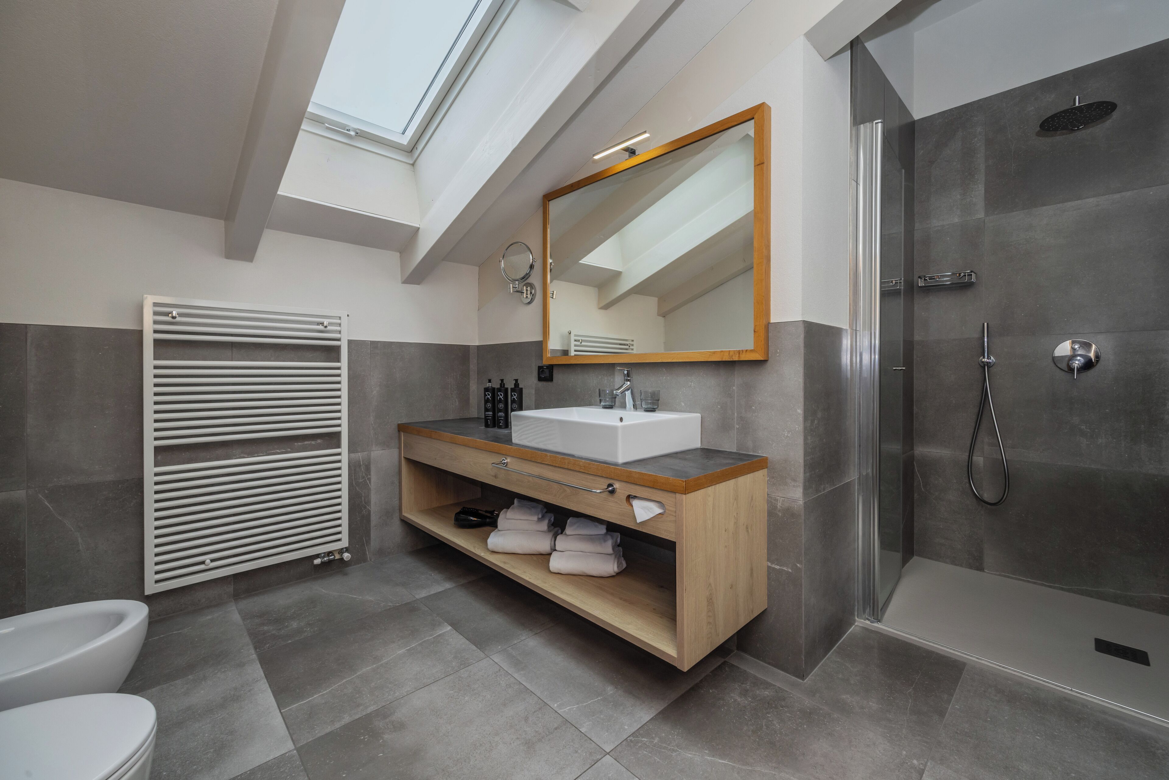 A modern bathroom with a skylight, a large mirror, a shower, and a vanity with towels.