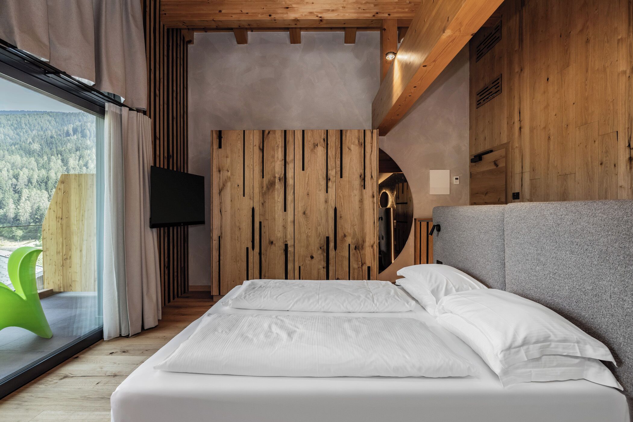 A bedroom with a bed, a television, and a view of a forest through a window.