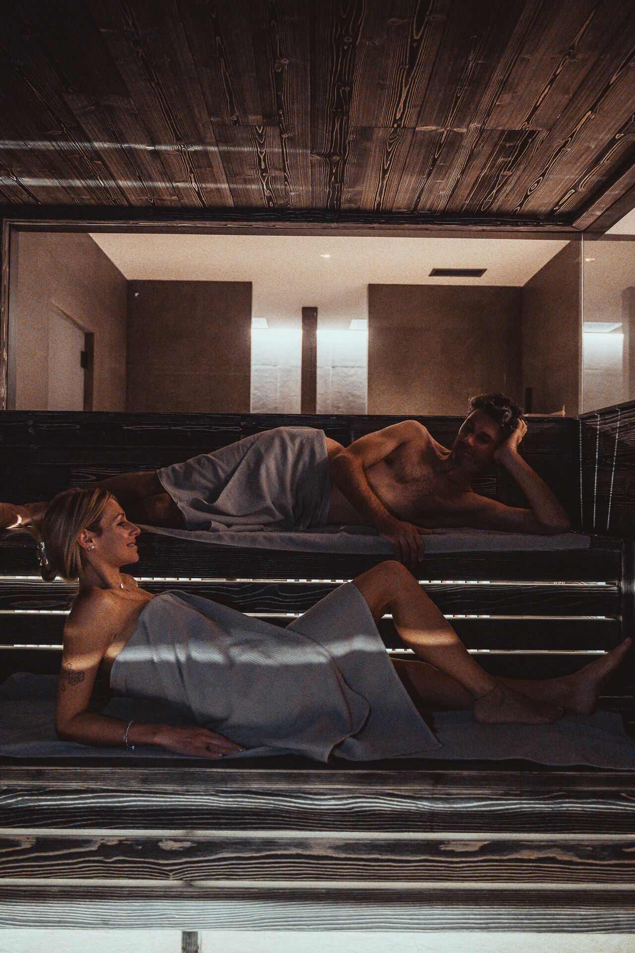 A room with a wooden ceiling and a large window. There are two people lying on a bed.