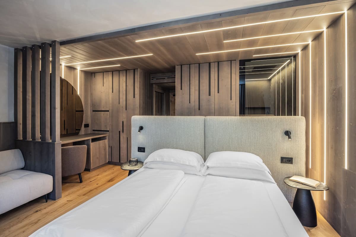 Une chambre d’hôtel moderne avec un grand lit, un bureau et une chaise. On y trouve une table ronde sur laquelle sont posés une lampe et un chapeau. La chambre est dotée de lambris en bois et d’un plafond éclairé par des luminaires linéaires.