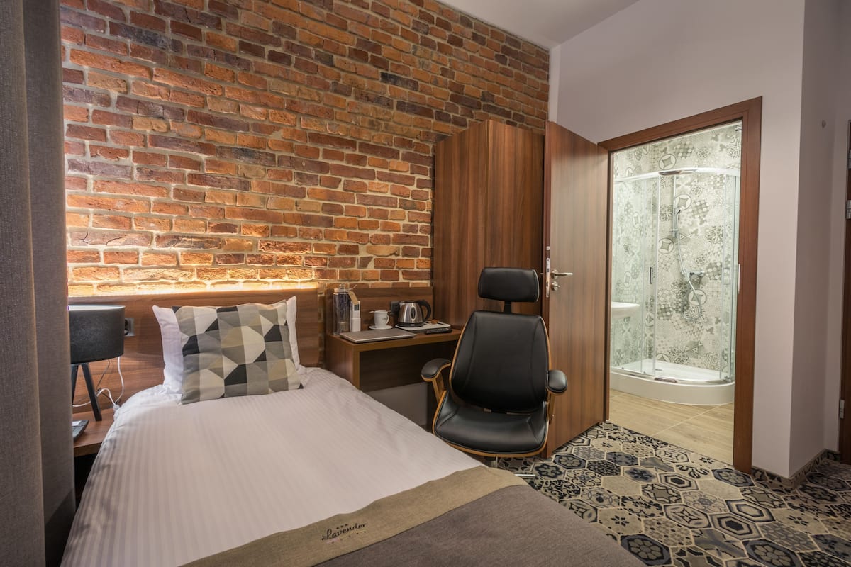 Habitación de hotel con pared de ladrillo, cama con almohada geométrica, escritorio con hervidor, silla negra y puerta de ducha de vidrio.