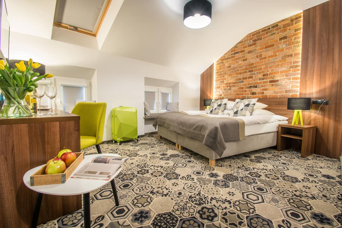 Una habitación de hotel moderna con pared de ladrillo decorativa, piso estampado, cama con almohadas, mesita de noche con lámpara, silla, escritorio con un jarrón de flores y ventana con cortinas.