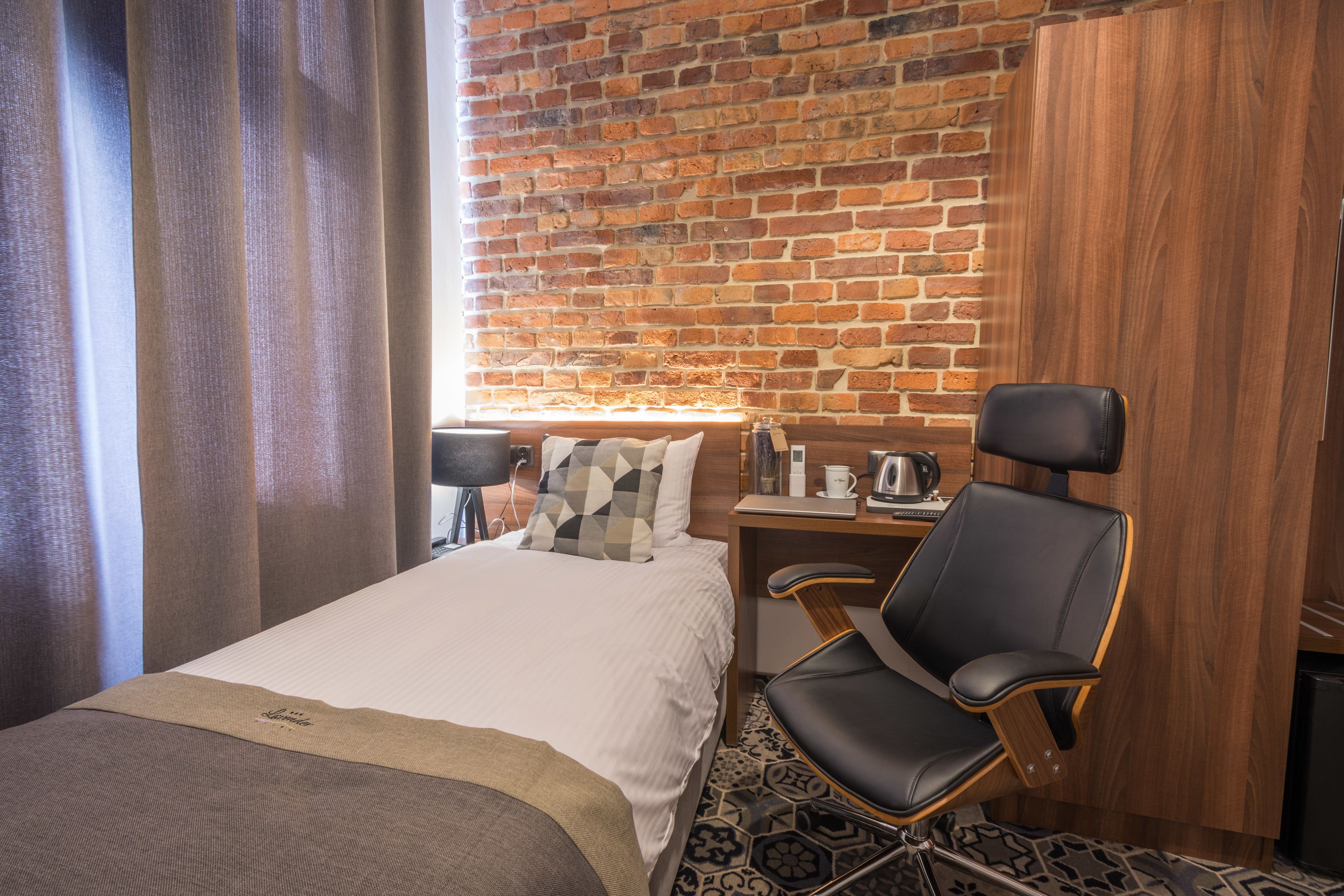 A hotel room with a brick wall, a bed with a white comforter, a black leather chair, a wooden desk, and a window with curtains.