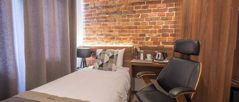 A hotel room with a brick wall, a bed with a white comforter, a black leather chair, a wooden desk, and a window with curtains.