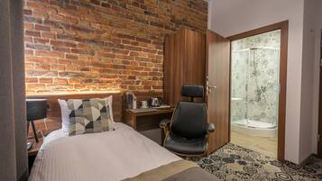 Habitación de hotel con pared de ladrillo, cama con almohada de estampado geométrico, escritorio con una computadora portátil, una silla y zona de ducha con azulejos estampados.