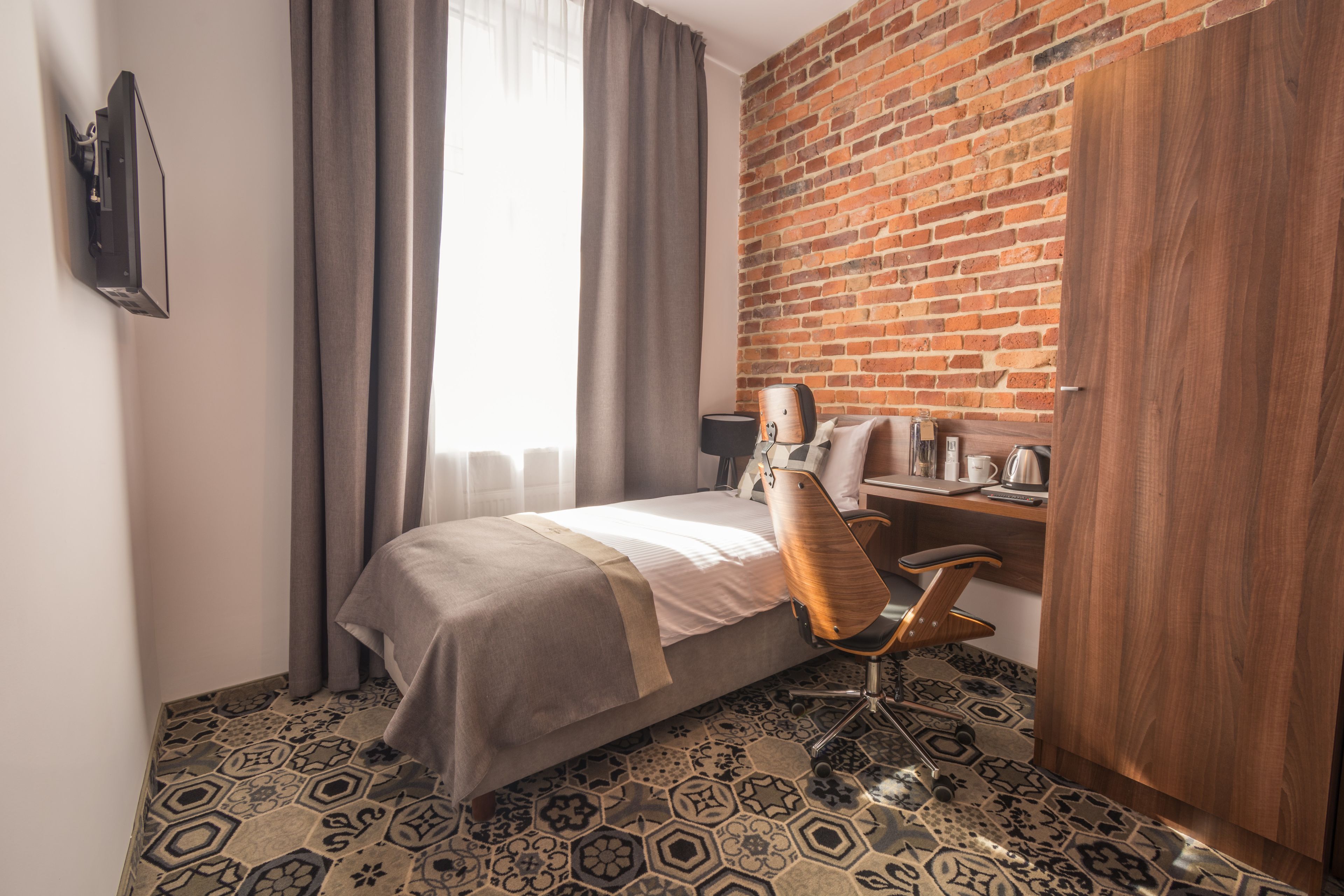 A hotel room with a bed, a desk, a chair, a wardrobe, and a brick wall.