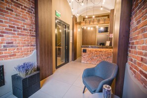 A reception area with a brick wall, a reception desk, a grey chair, and a small planter with flowers. - Hotel Lavender (Kraków)