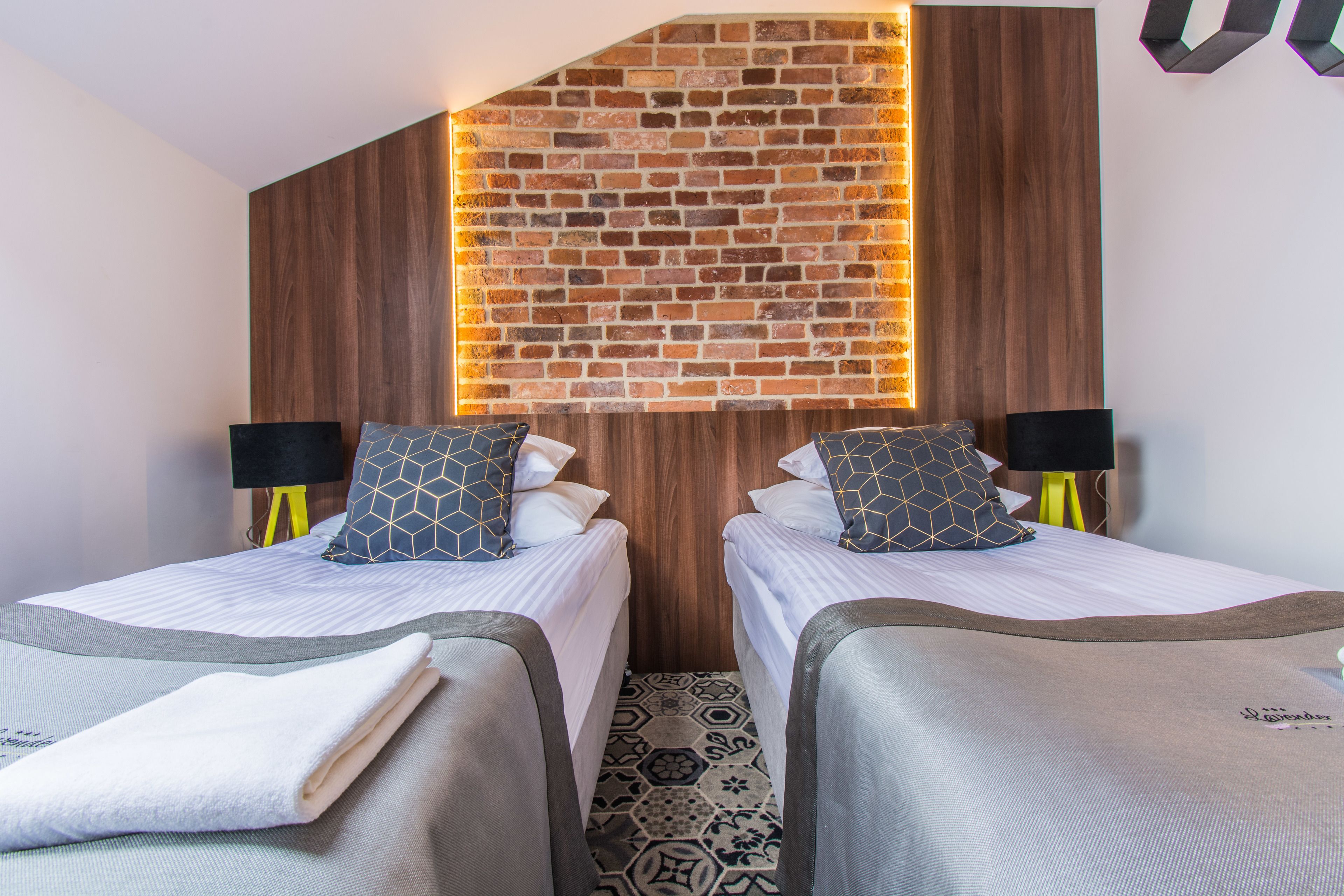 Ein Hotelzimmer mit zwei Betten, jeweils mit Kissen und Lampe. Das Zimmer hat eine Ziegelwand mit einer Holzvertäfelung und einen gemusterten Boden.