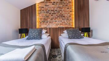 A hotel room with two beds, each with a pillow and a lamp. The room has a brick wall with a wooden panel and a patterned floor.