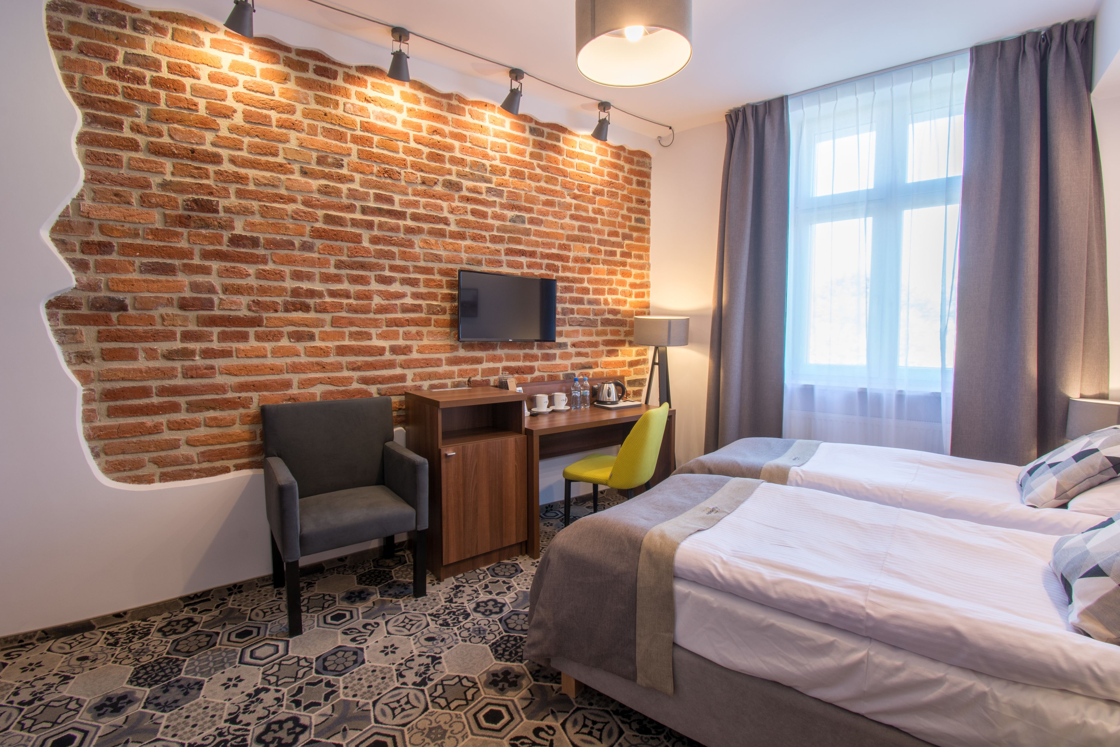 A hotel room with a brick wall, a desk with a chair, a bed, and a window with curtains.
