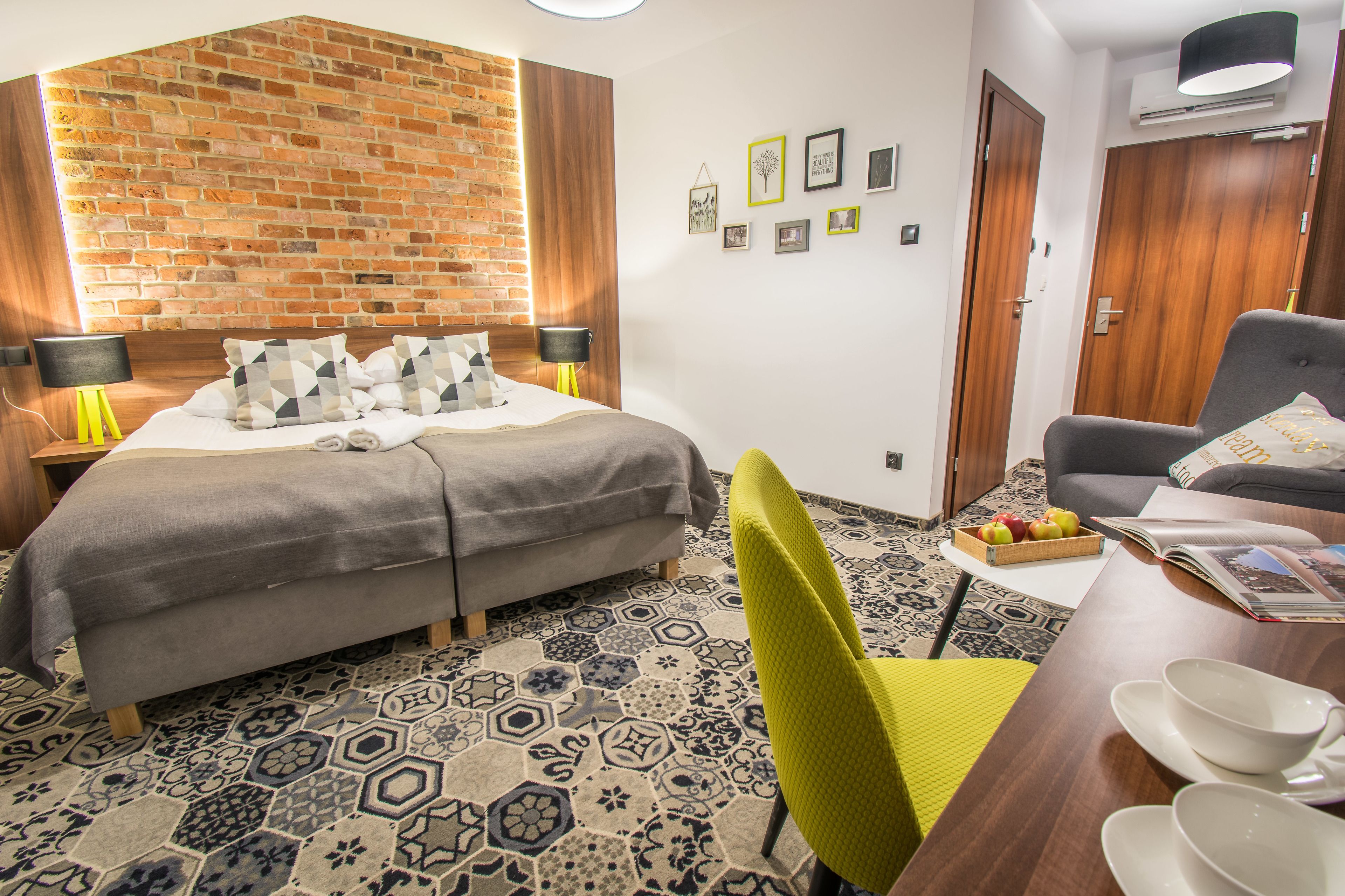 Una habitación de hotel moderna con una pared de ladrillo decorativa, una cama bien tendida, una zona de comedor con mesa y sillas, y una pequeña cesta de frutas.