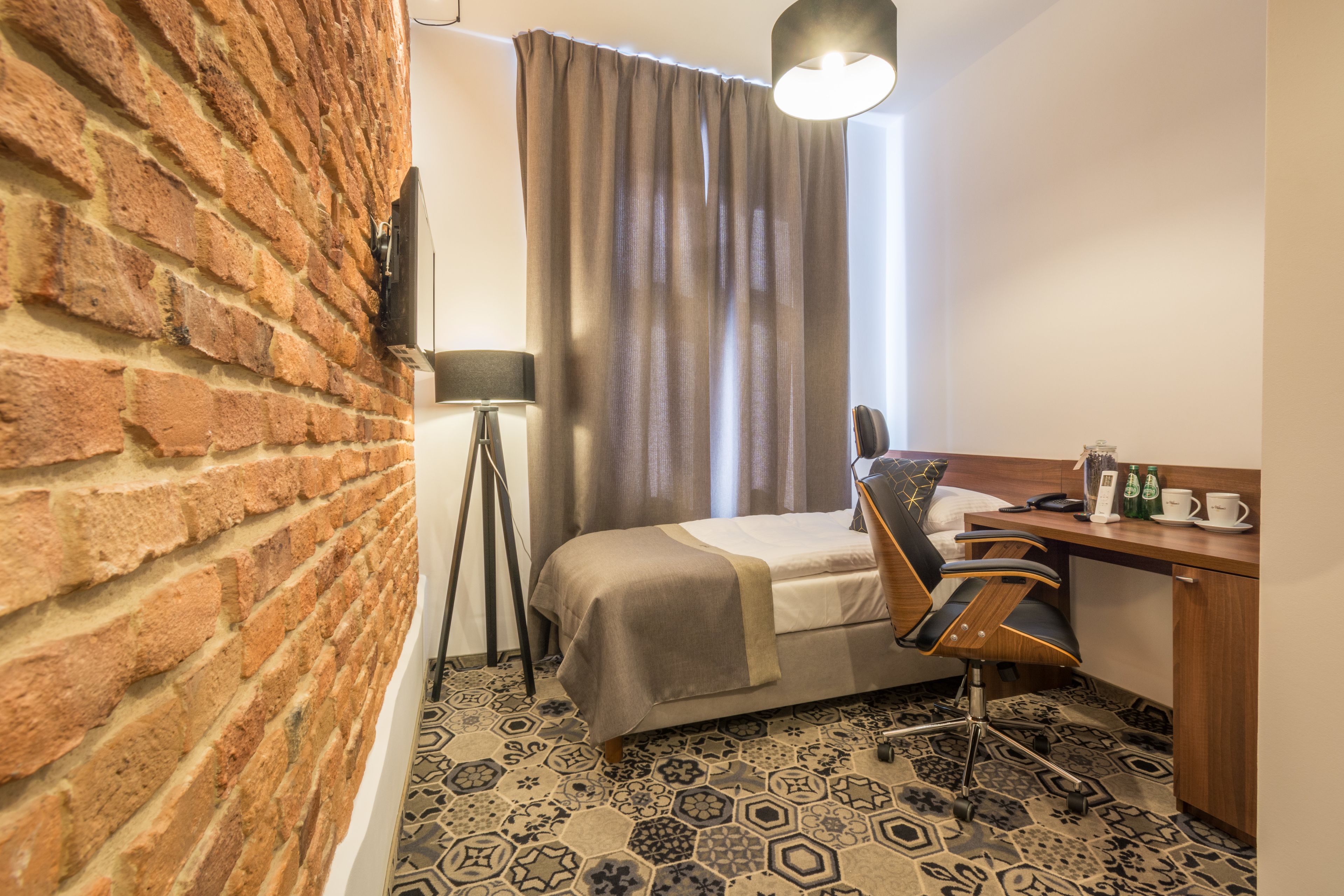 A hotel room with a brick wall, a bed with a headboard, a desk with a chair, a lamp, and a patterned floor.