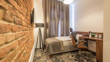 A hotel room with a brick wall, a bed with a headboard, a desk with a chair, a lamp, and a patterned floor.