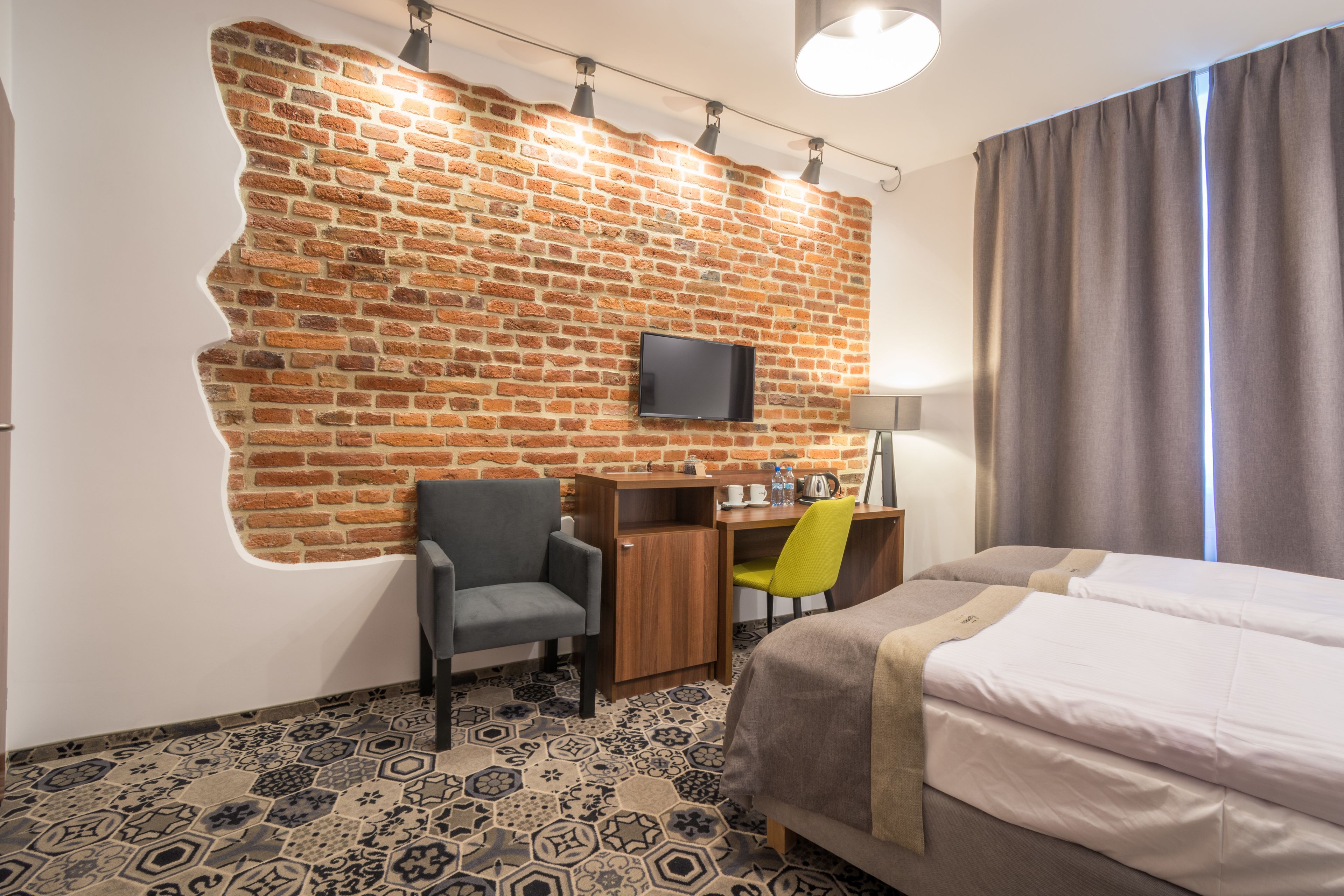 Hotellihuone, jossa on tiiliseinä, taulutelevisio, työpöytä tuolilla, sänky ja kuviollinen lattia.