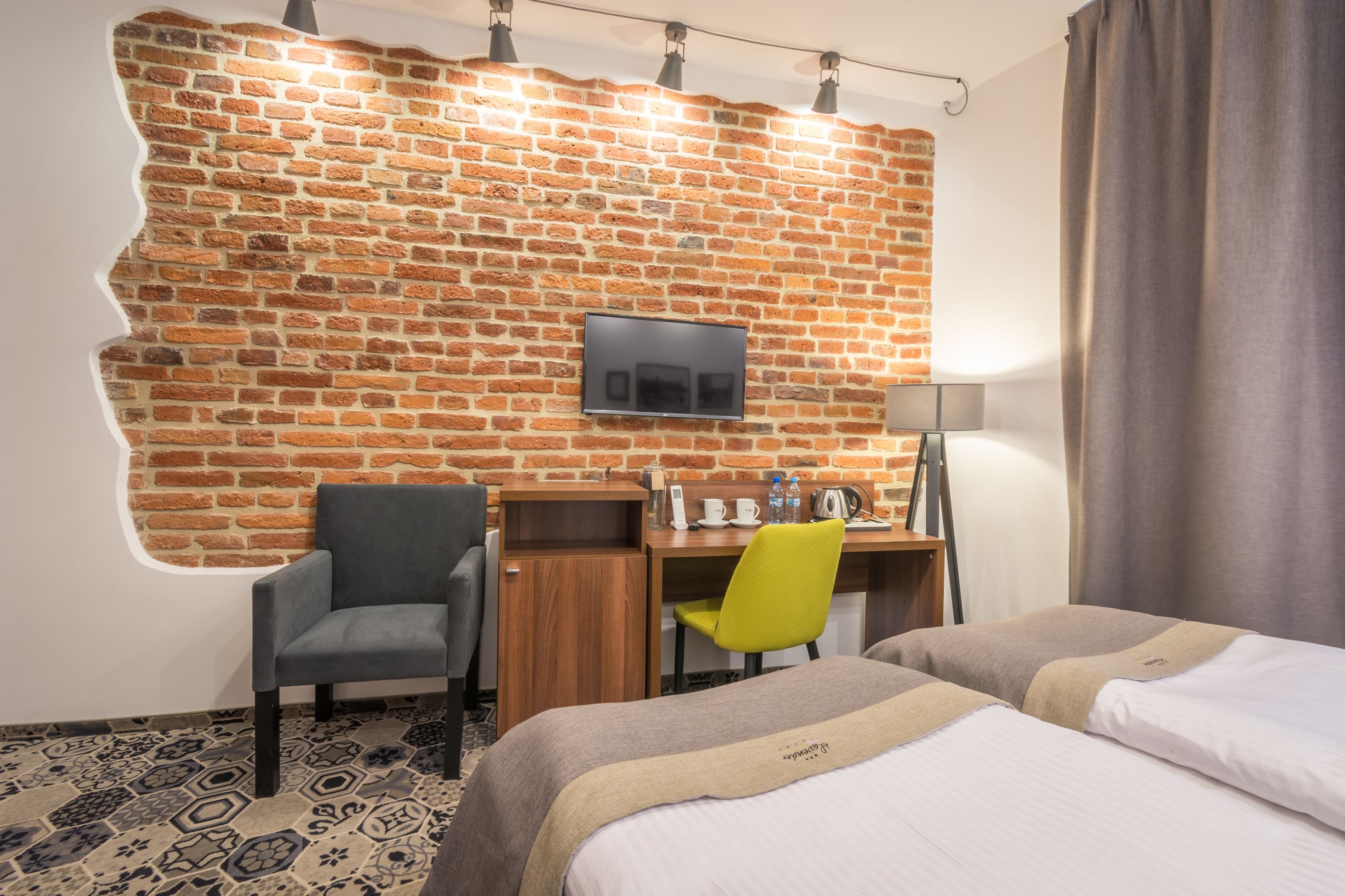A hotel room with a brick wall, a flat-screen TV, a desk with a chair, a bed, and a lamp.