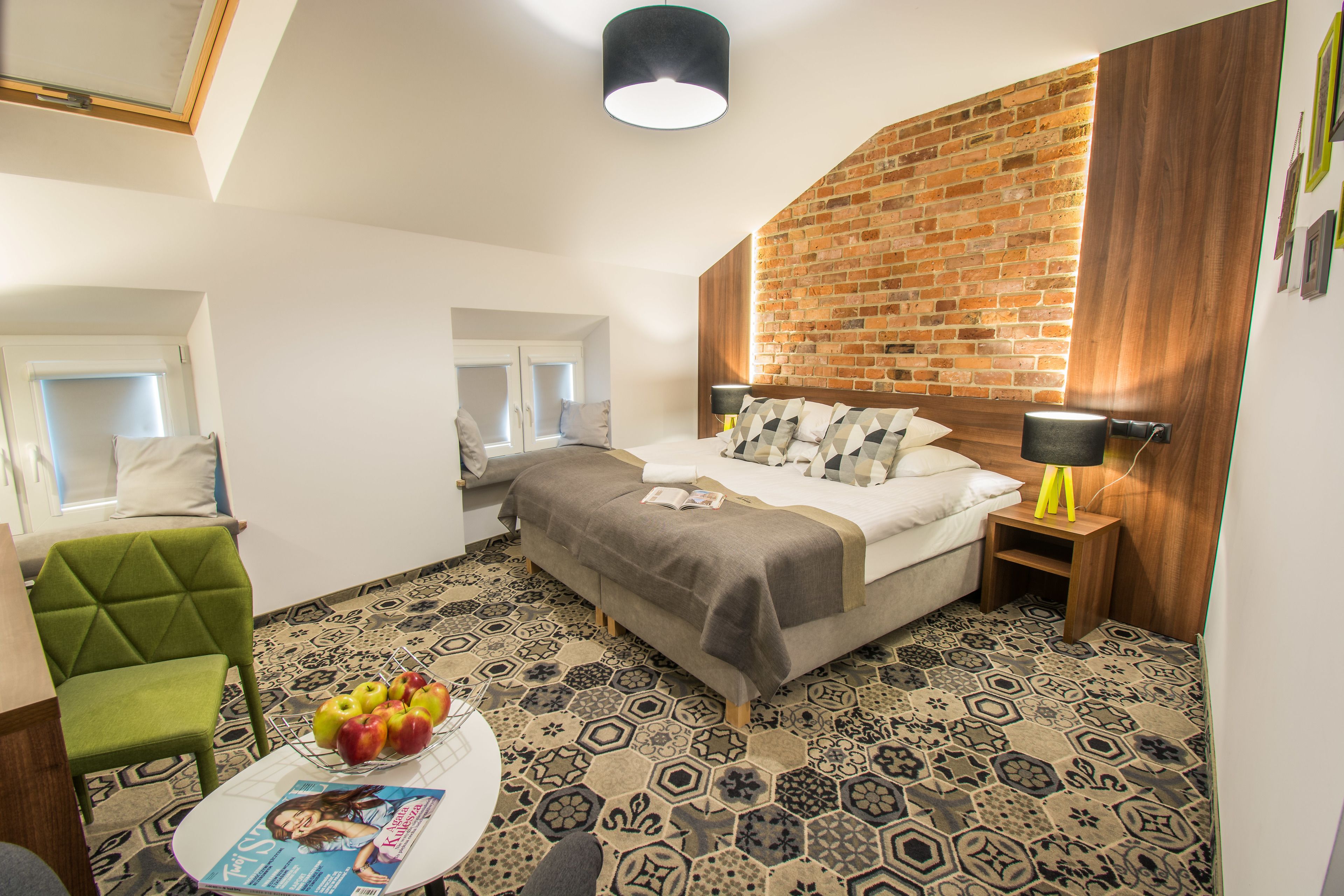 Habitación de hotel con pared de ladrillo, cama con cabecera de madera, un sillón verde, una mesa redonda con una revista y piso estampado.