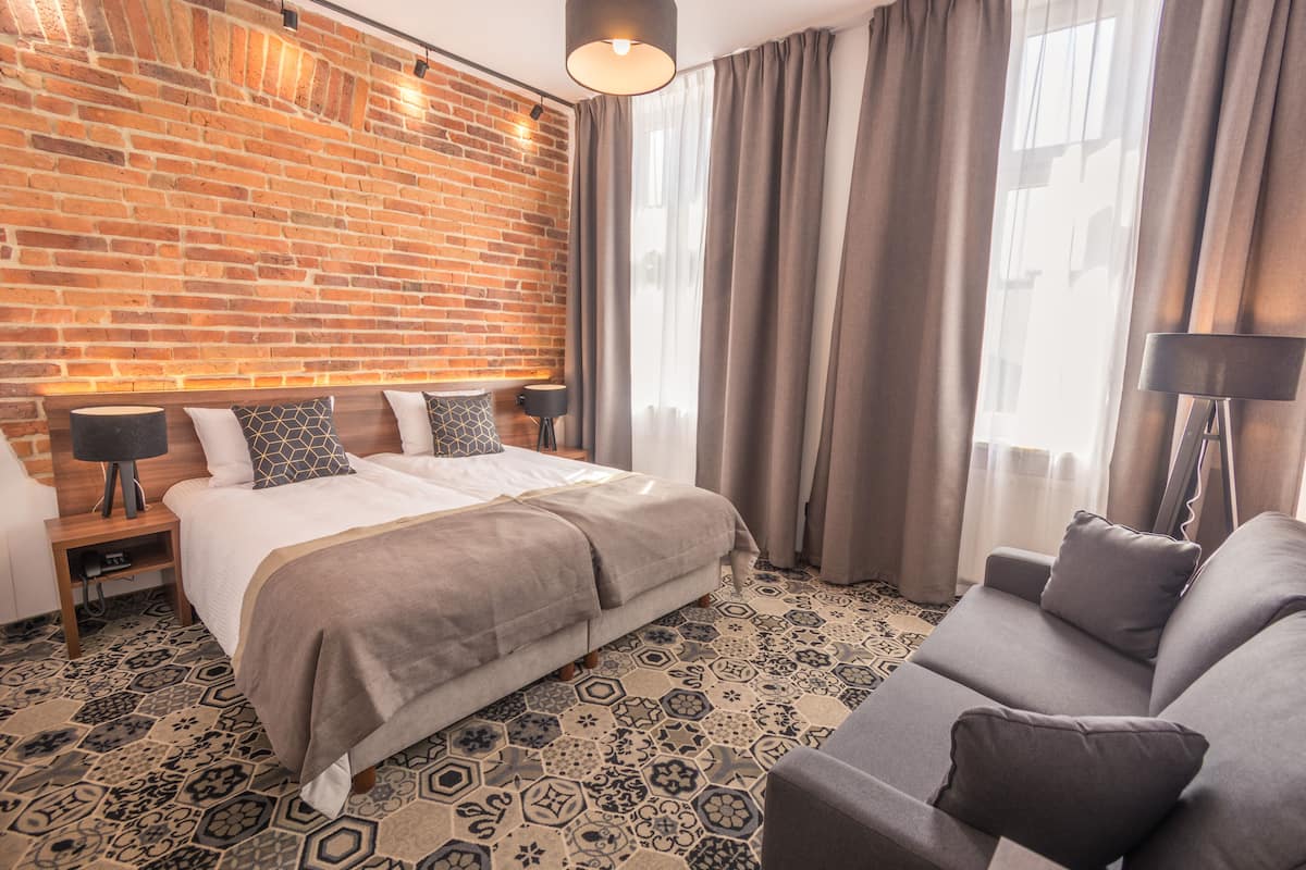Habitación de hotel con pared de ladrillo, cama con cubrecama gris, sofá gris y alfombra estampada.