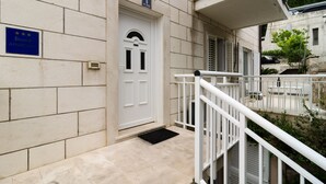 A building entrance with a white door, a blue sign indicating an apartment, and a small outdoor seating area with a white railing. - Apartments Carmelitta (Dubrovnik)