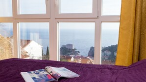 A bed with a purple blanket and a magazine, a window with a yellow curtain, and a view of a coastal town and sea. - Apartments Carmelitta (Dubrovnik)