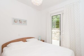 A bedroom with a bed, a nightstand, a wall-mounted light, and a window with a view of greenery. - Apartments Carmelitta (Dubrovnik)