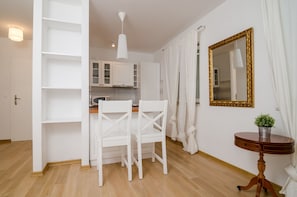 A modern kitchen with white cabinets, a wooden dining table with chairs, a pendant light, and a round wooden side table with a plant. - Apartments Carmelitta (Dubrovnik)
