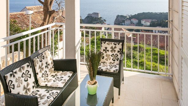 A balcony with wicker furniture, a potted plant, and a view of the sea and buildings. - Apartments Carmelitta (Dubrovnik)