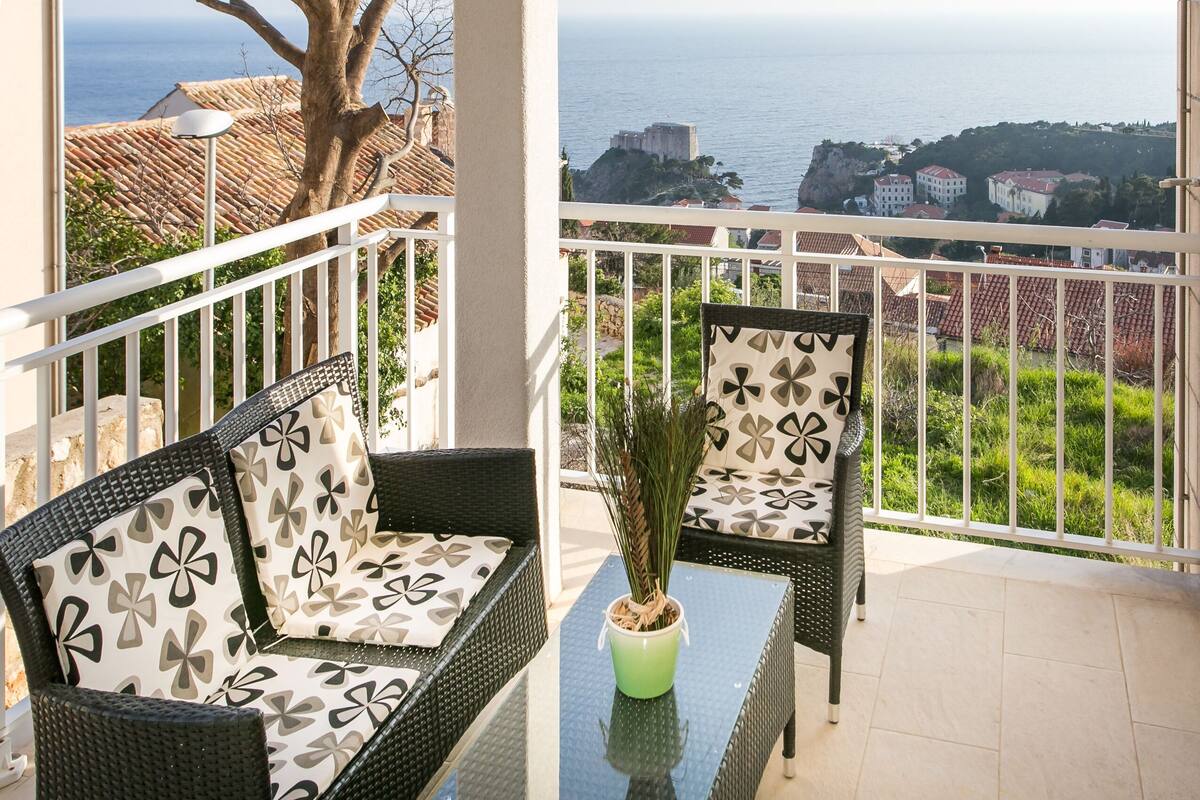 A balcony with wicker furniture, a potted plant, and a view of the sea and buildings.