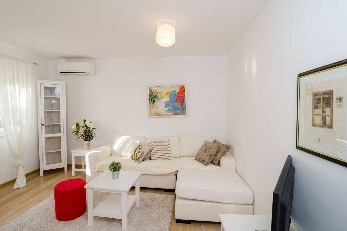 A modern living room with a white sofa, a small table, a red ottoman, a TV, and a painting on the wall.