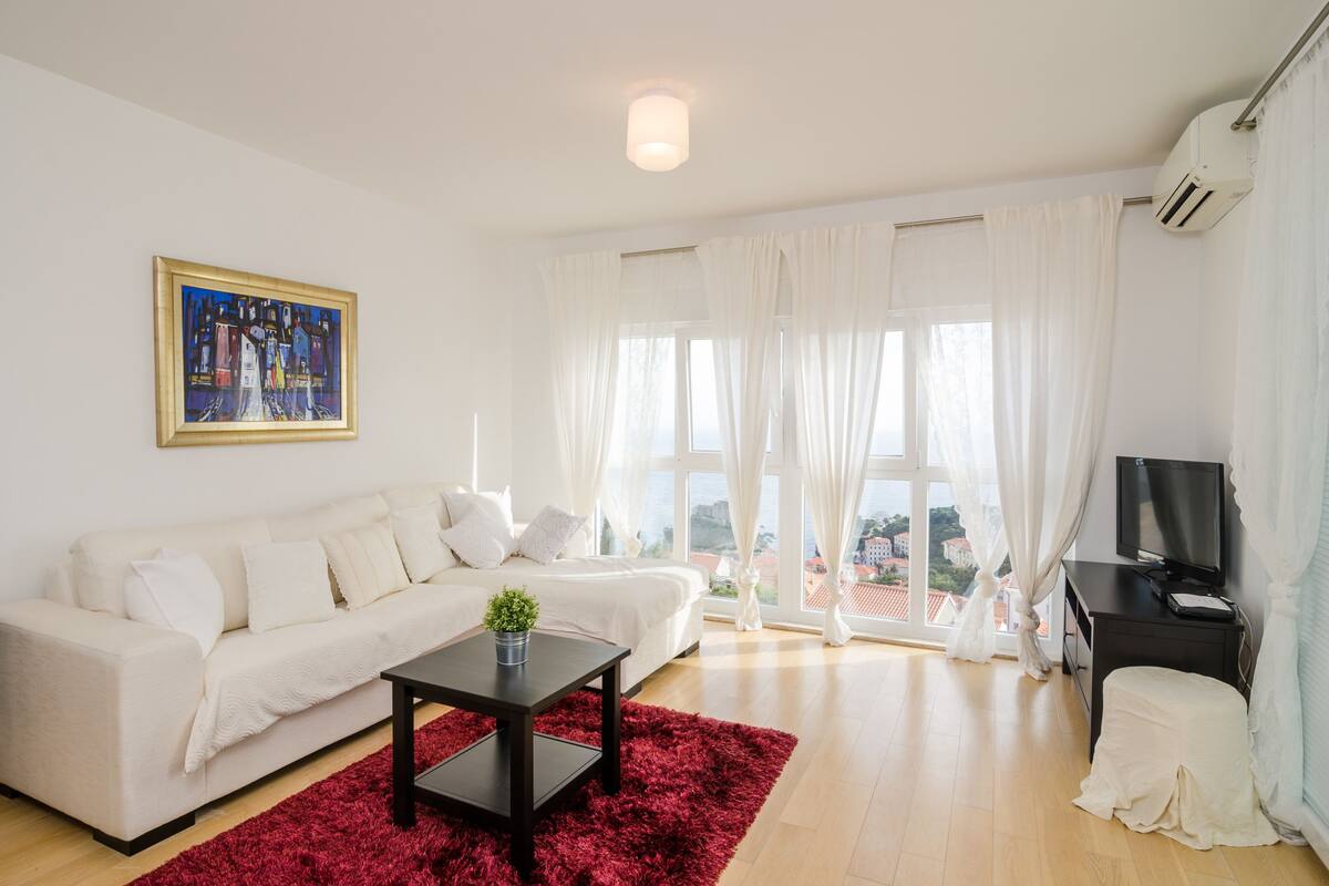 A modern living room with a white sofa, a red rug, a painting on the wall, and a TV on a black stand.