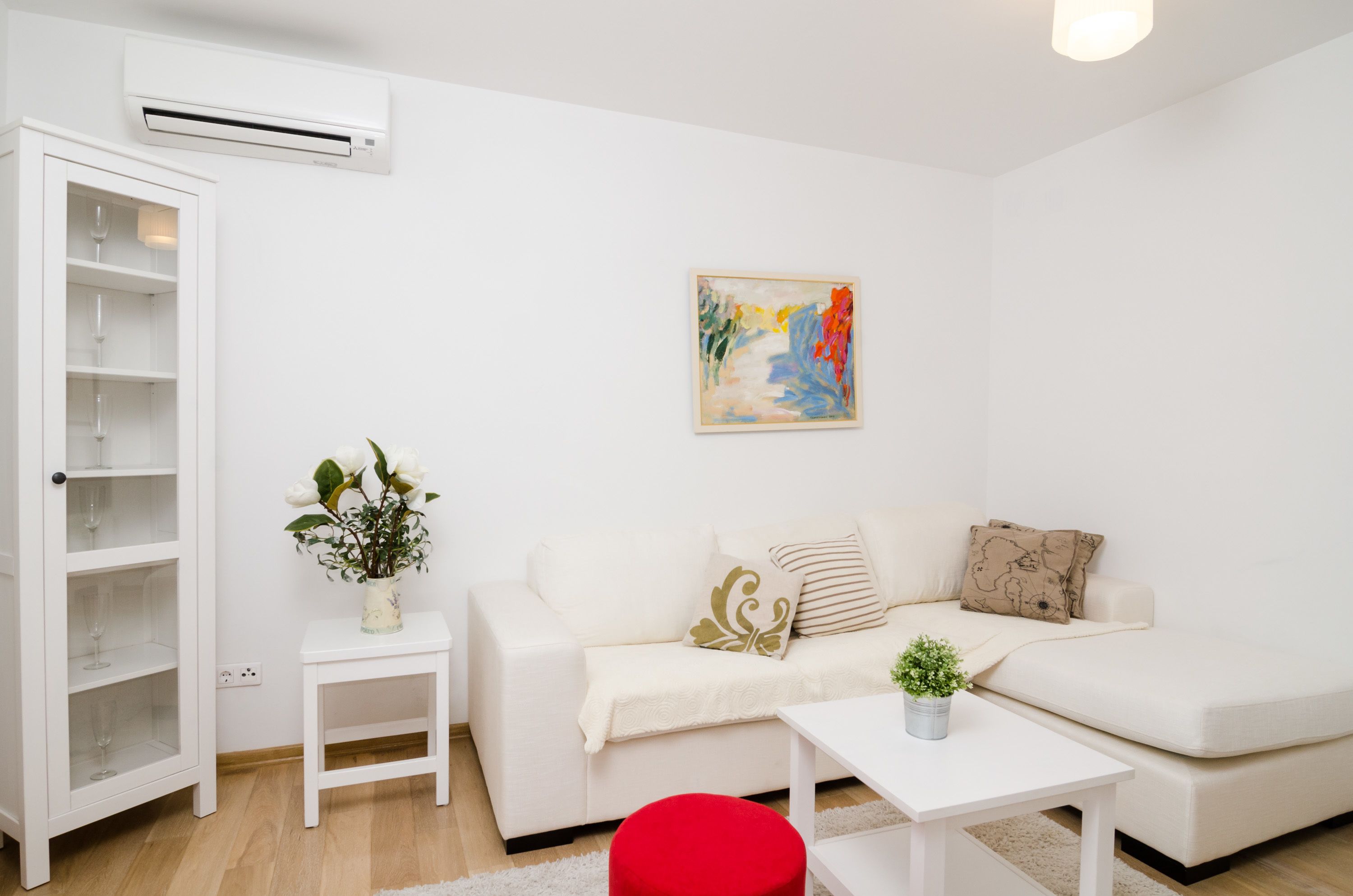 En moderne stue med en hvid sofa, et lille sofabord med en vase blomster, en rød fodskammel og et billede på væggen.