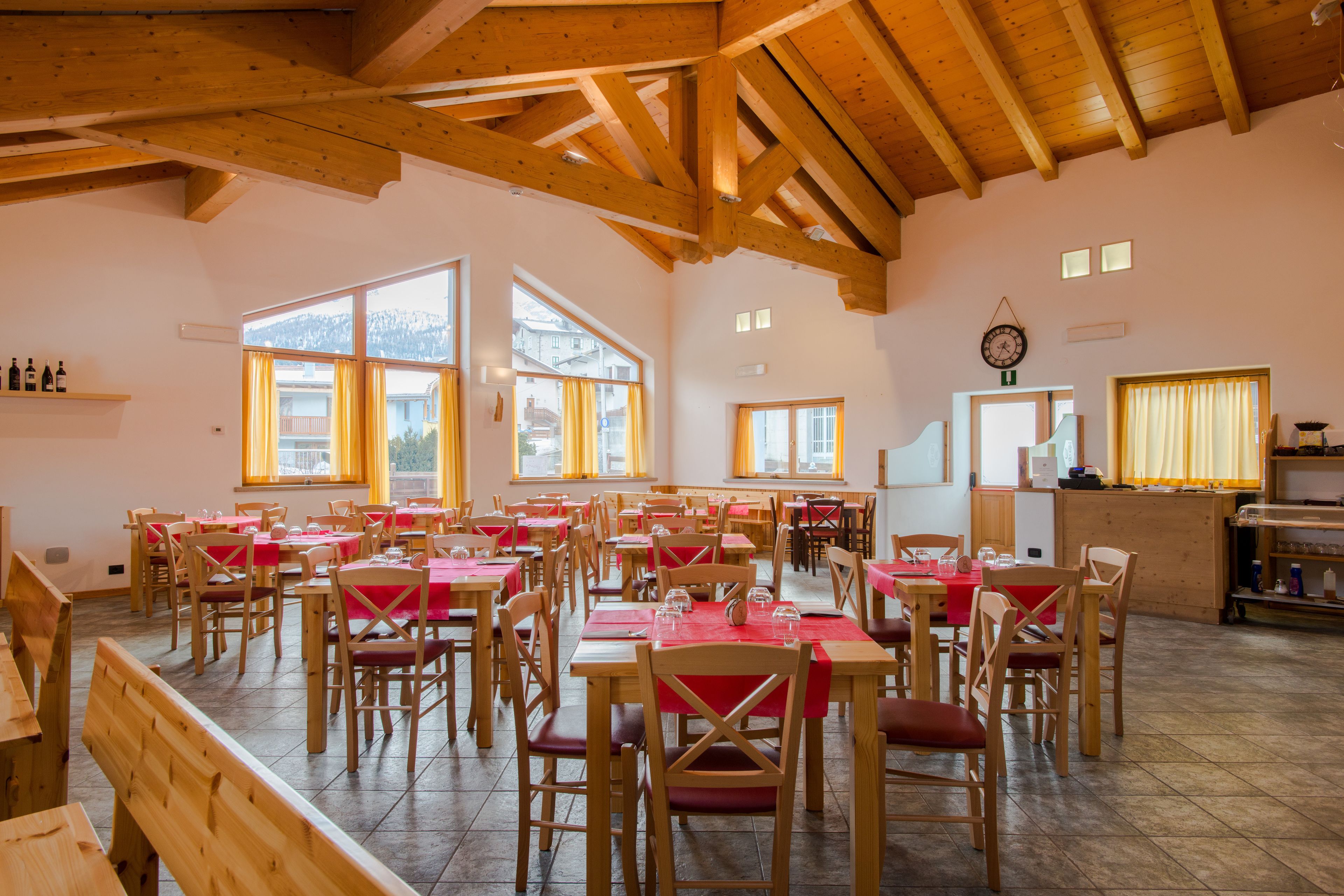 A dining area with multiple tables and chairs, wooden beams on the ceiling, and large windows allowing natural light.