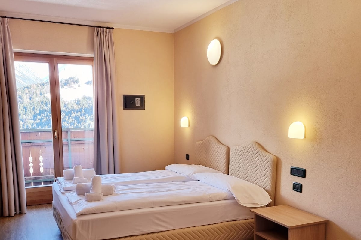 Habitación de hotel con una cama grande, dos mesitas de noche, suelo de madera y vistas a un paisaje montañoso a través de una ventana con cortinas.