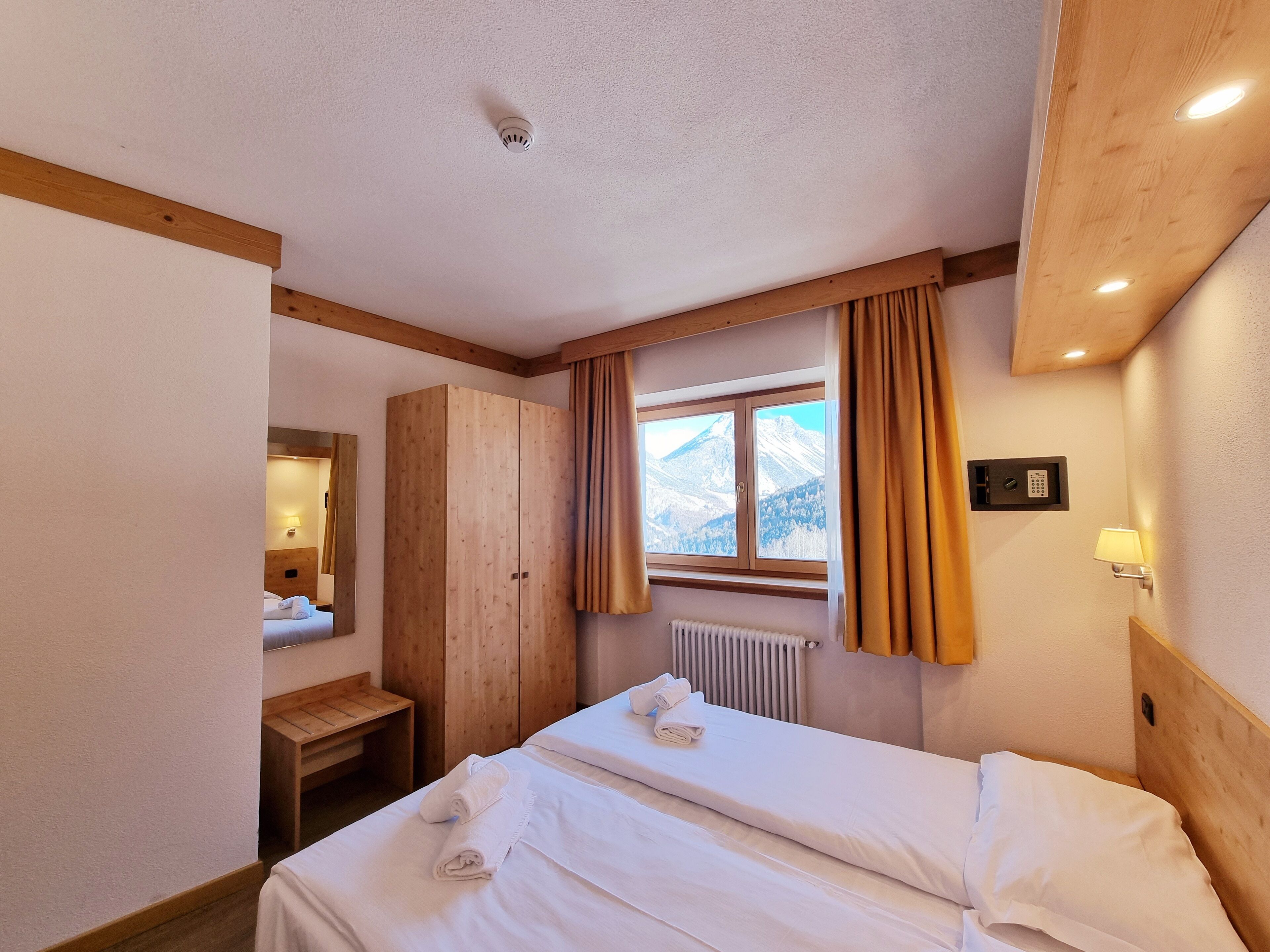 Habitación de hotel con cama, armario de madera, ventana con cortinas, radiador y lámpara fijada a la pared.