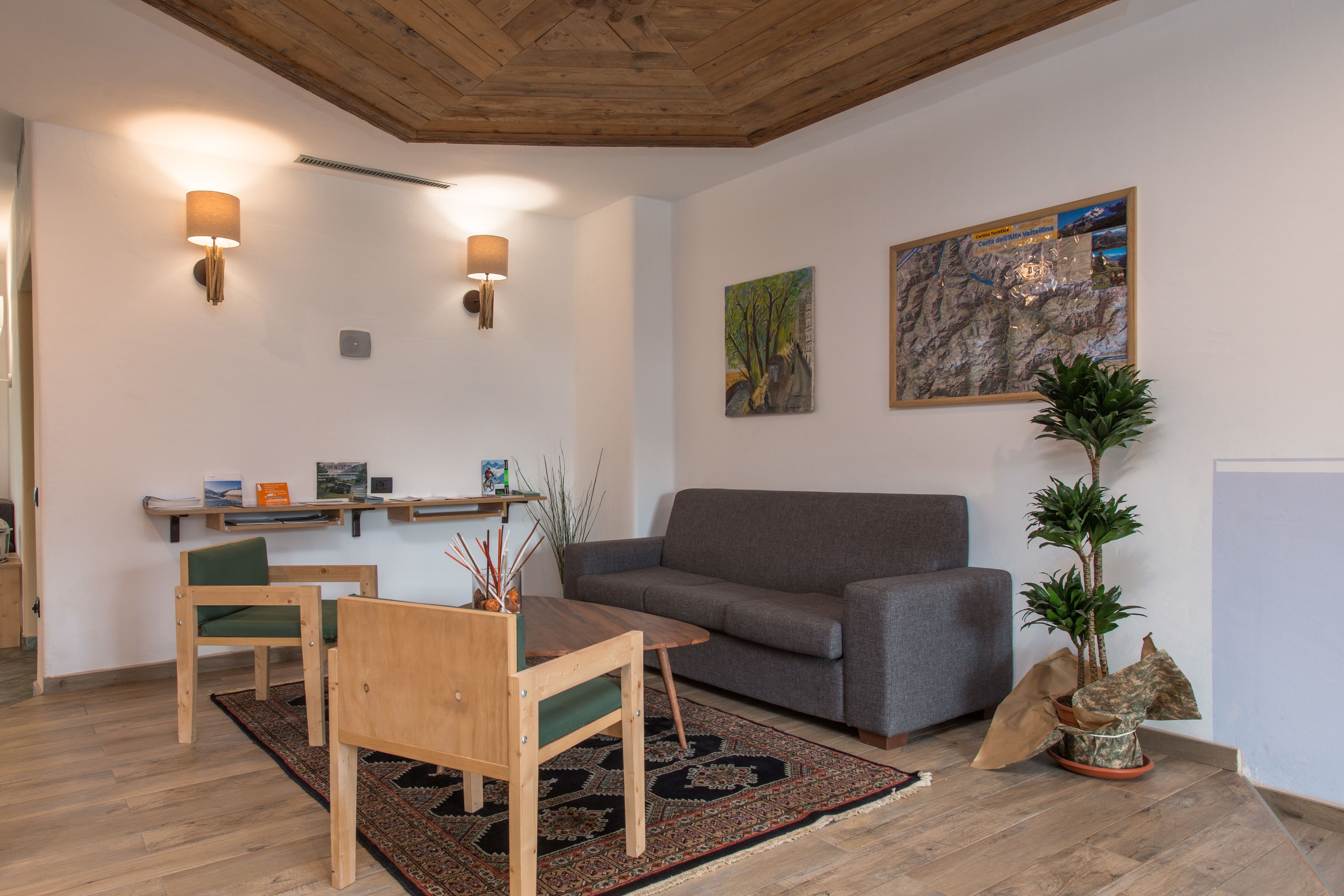 A living room with a wooden ceiling, a grey sofa, a wooden coffee table, and a green chair. There is a small shelf with books and decorative items, and a painting on the wall.