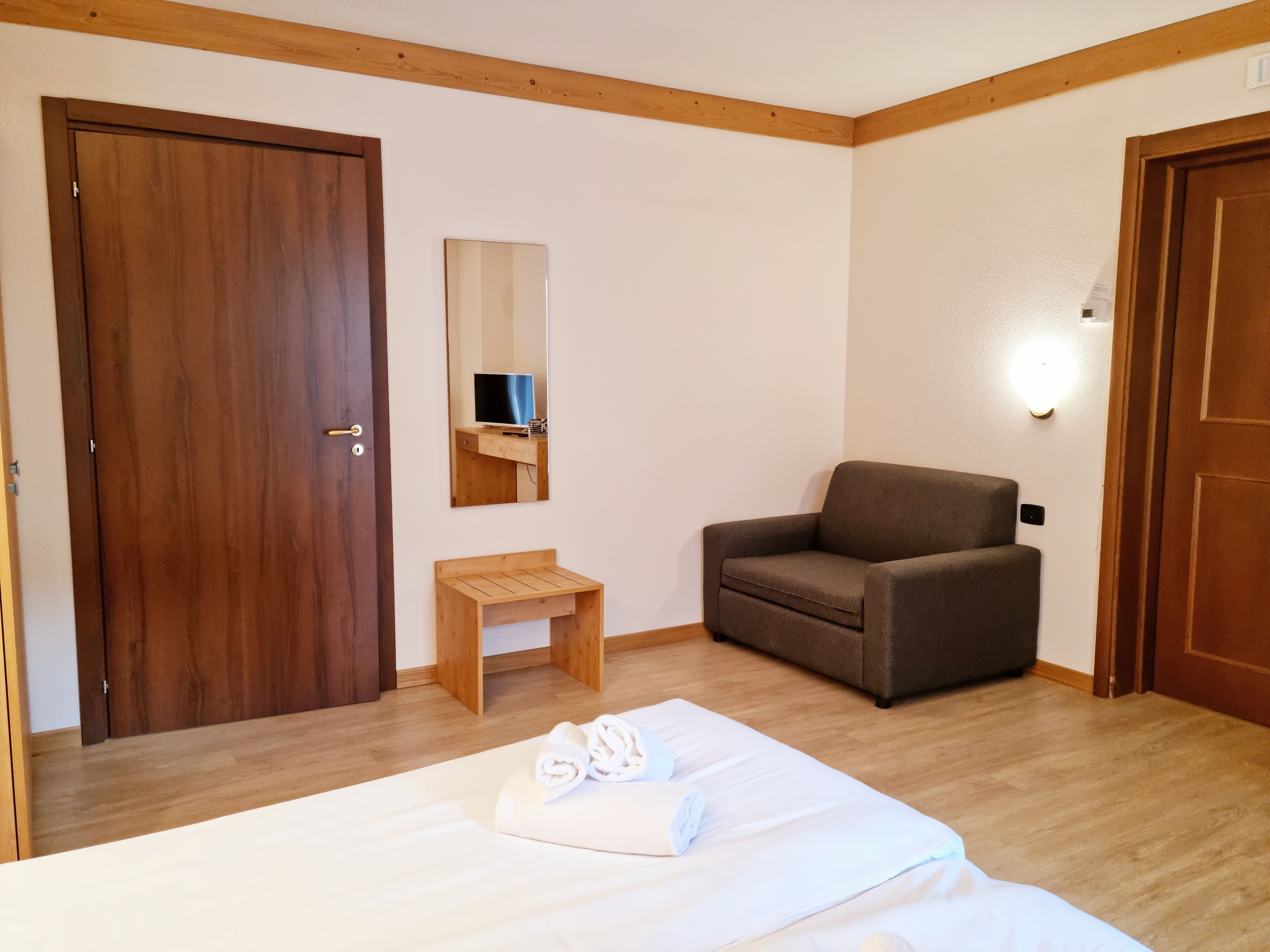 A hotel room with a bed, a wooden chair, a small wooden table, a mirror, and a wall-mounted TV.