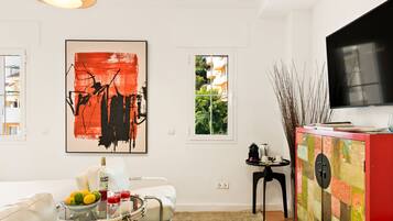 A modern living room with a white sofa, a coffee table with drinks, a red cabinet with a TV, and a large abstract painting on the wall.