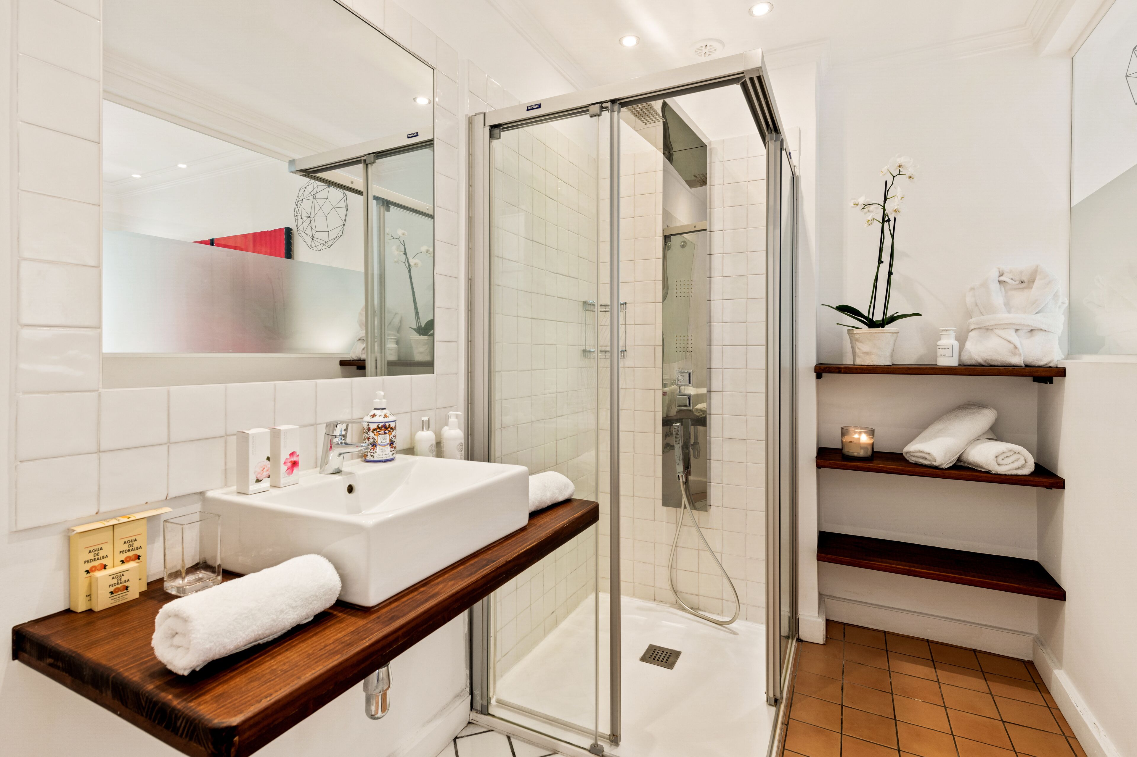 A modern bathroom with a glass-enclosed shower, a white sink, and wooden shelves holding towels and toiletries.
