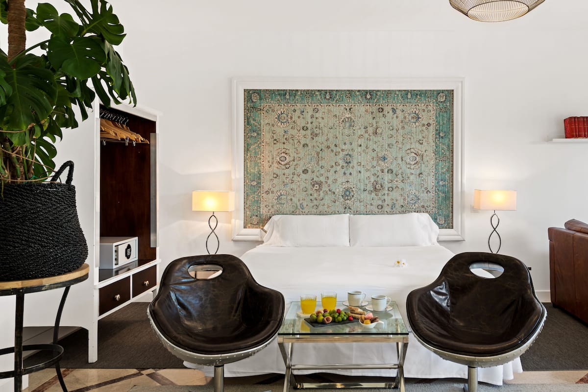 Une chambre d’hôtel avec un grand lit, deux fauteuils, une petite table avec le petit-déjeuner, et une grande tapisserie au mur.