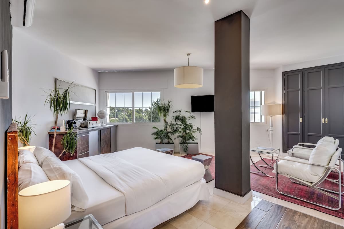 Une chambre moderne avec un grand lit, un bureau et une chaise. On y trouve plusieurs plantes et un téléviseur fixé au mur.