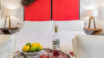 A hotel room with a red accent wall, a bed with white linens, a bedside table with a bottle of liquor, glasses, and a bowl of fruit.