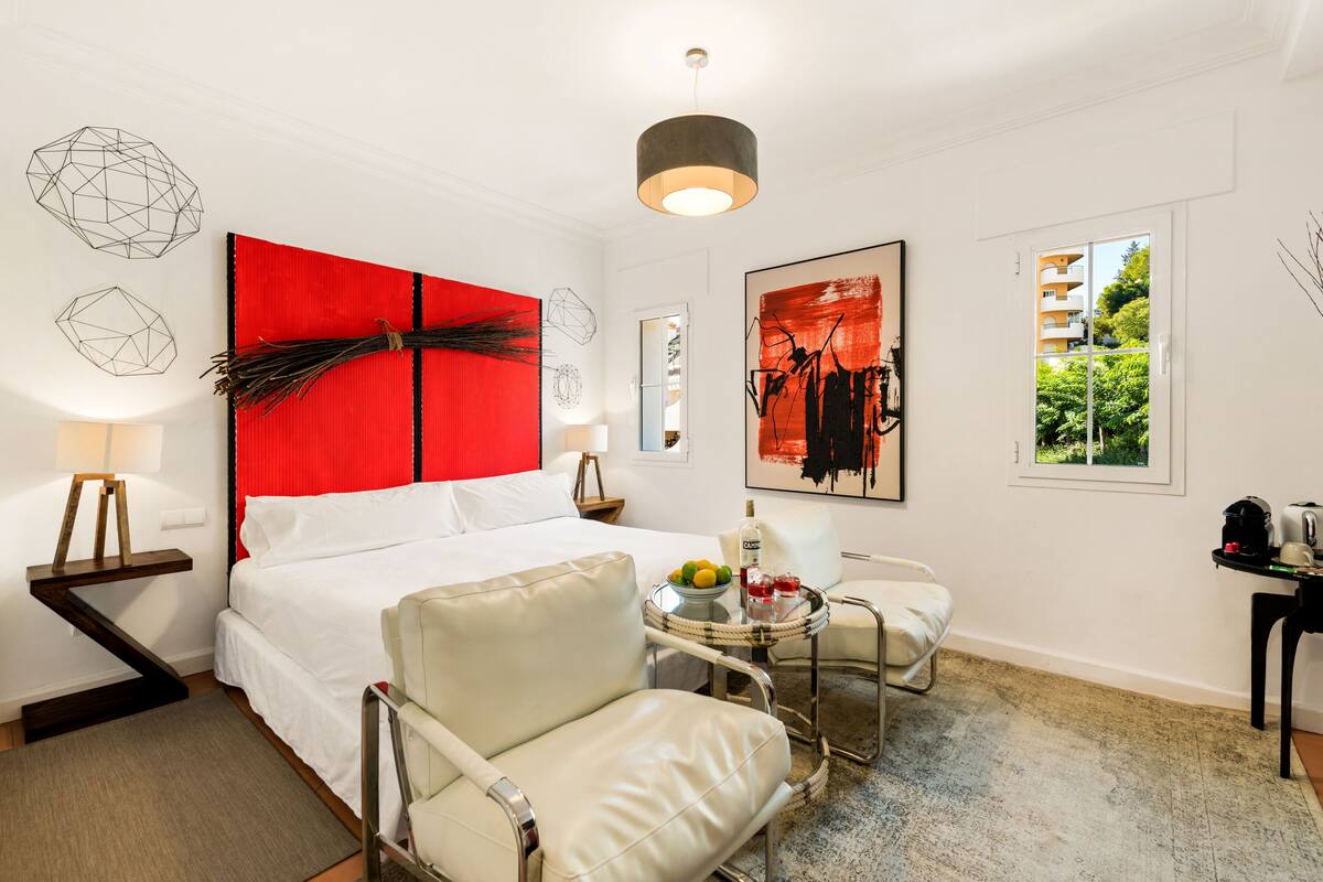 Une chambre d’hôtel moderne avec un grand lit, un mur d’accent rouge, un coin repas avec une petite table et des chaises, et des œuvres d’art accrochées aux murs.