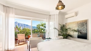 A hotel room with a large bed, a desk, and a chair. There is a view of mountains and a balcony with plants.